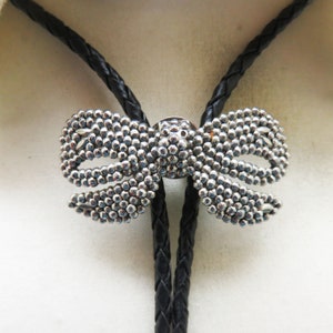 May include: A silver bow tie bolo tie with a black braided cord. The bow tie is made of small silver beads.