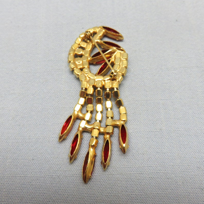 Pode incluir: Um broche dourado com um design de strass vermelho. O broche tem a forma de uma l&aacute;grima com uma franja de strass vermelhos pendurados.