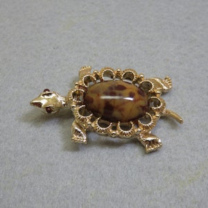 May include: Gold-tone turtle brooch with a brown and white marbled stone cabochon set in a filigree design.