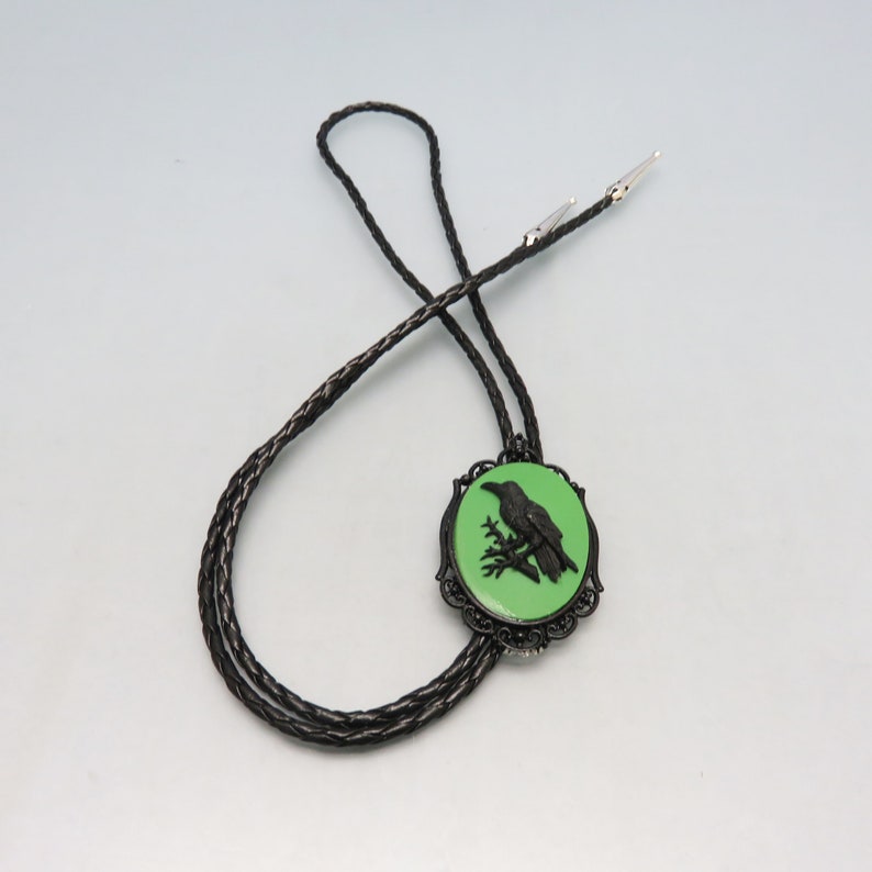 K&ouml;nnte beinhalten: Schwarze Bolo-Krawatte mit einem gr&uuml;nen Kamee, das eine schwarze Silhouette eines Rabens auf einem Ast zeigt.