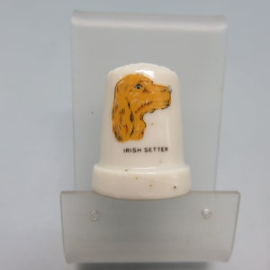 May include: White ceramic thimble with a hand-painted image of an Irish Setter dog's head. The dog is brown and white and the text "IRISH SETTER" is printed below the image.