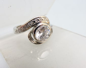 May include: A silver ring with a large, clear, round gemstone surrounded by smaller, clear gemstones. The band is twisted and has a delicate design.