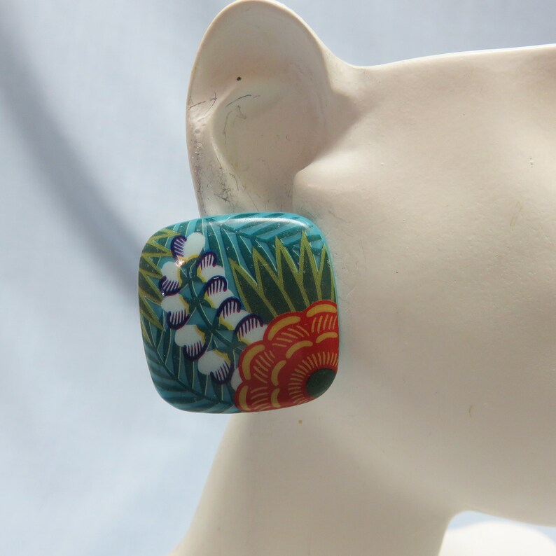 Op de afbeelding: Vierkante oorbellen met een bloemenmotief. De oorbellen zijn turquoiseblauw met groene, witte, oranje en rode bloemen.