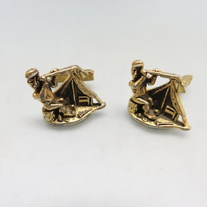 May include: A pair of gold-tone cufflinks featuring a detailed design of a person sitting by a tent with an axe.