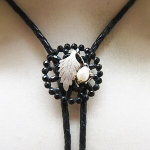 以下が含まれることがあります： 黒いボーラタイ。中央に黒いビーズを使った円形デザイン。銀色の葉と白い真珠がアクセント。タイは黒い編み込みコードを使用しています。