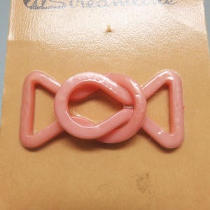 May include: A light pink plastic buckle with a central circular loop and two triangular side loops, attached to a tan cardboard backing card with the partial text "Streamline" printed in dark blue at the top of the card.