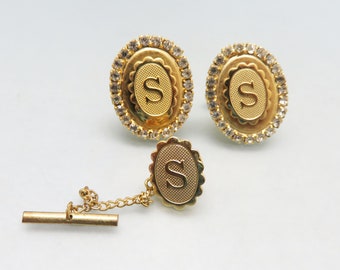 May include: A set of gold cufflinks and a tie tack featuring an oval design with a clear rhinestone border and the letter "S" in the center.