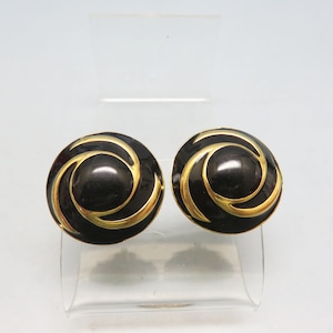 May include: Pair of black and gold-tone swirl design clip-on earrings. The earrings are round with a glossy black center and a gold-tone swirl pattern around the edge. The earrings are displayed on a clear stand.