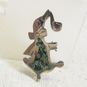 May include: A silver and abalone shell pendant in the shape of a stylized animal with a raised ear and a curved horn. The pendant is on a white fabric background.