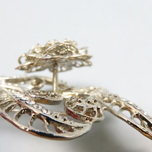 May include: Silver filigree brooch with a large, ornate, silver flower design. The flower has a raised center and is attached to the brooch with a pin.