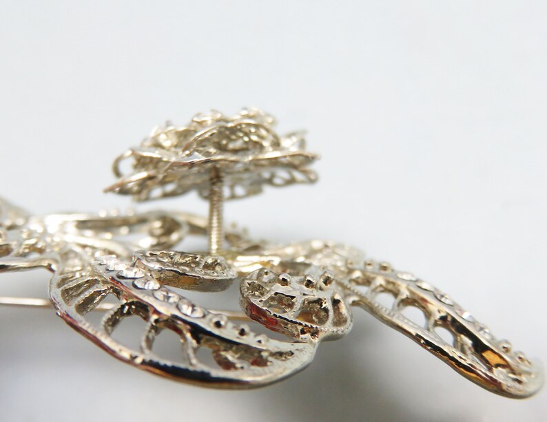 May include: Silver filigree brooch with a large, ornate, silver flower design. The flower has a raised center and is attached to the brooch with a pin.