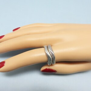 May include: A silver ring with a wavy, textured design. The ring is worn on a model's finger.