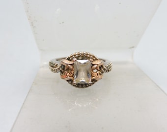 May include: A silver and rose gold ring with a clear square-shaped gemstone set in a decorative filigree design.