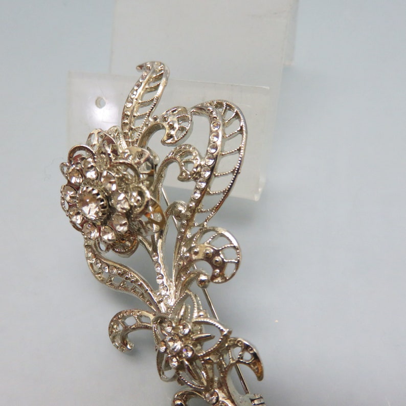 May include: A silver-toned brooch with a floral design. The brooch is adorned with clear rhinestones and has a delicate, intricate design.