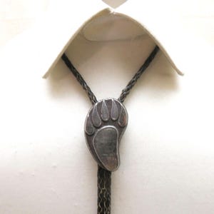 May include: A silver-toned bolo tie featuring a bear paw design. The pendant is attached to a braided black cord, displayed against a white shirt.
