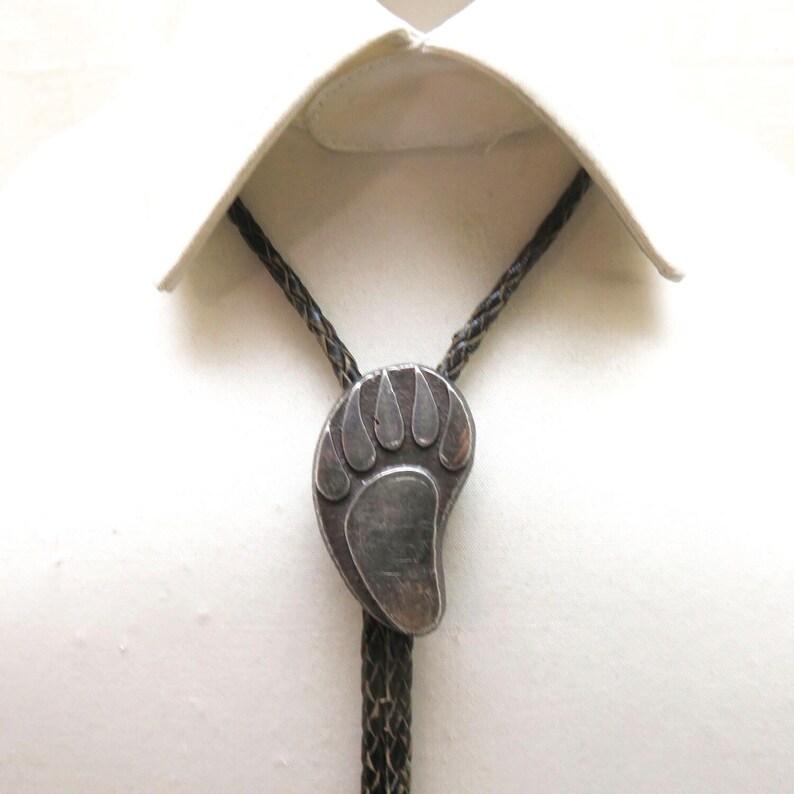 May include: A silver-toned bolo tie featuring a bear paw design. The pendant is attached to a braided black cord, displayed against a white shirt.