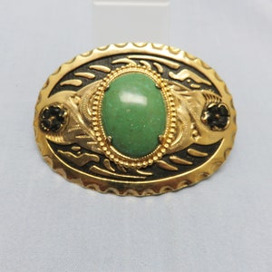 May include: A gold-tone oval brooch with a green cabochon stone set in a gold-tone bezel. The brooch has a black enamel design with floral accents.