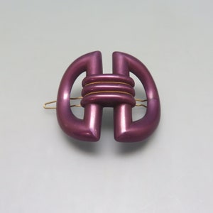 May include: A purple plastic hair clip with a gold accent. The clip is shaped like a figure eight with a knot in the center.