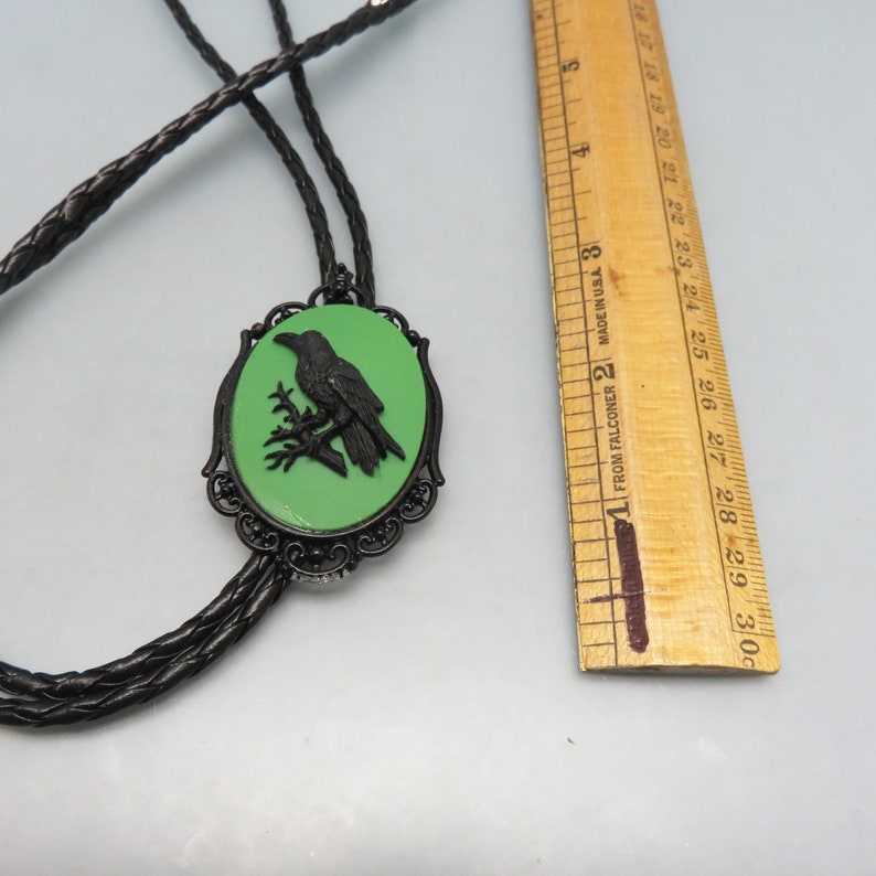 K&ouml;nnte beinhalten: Ein schwarzer Bolo-Krawattenhalter mit einem gr&uuml;nen Kamee, das eine schwarze Silhouette eines Rabens zeigt, der auf einem Ast sitzt. Das Kamee ist von einem schwarzen filigranen Rahmen umgeben.