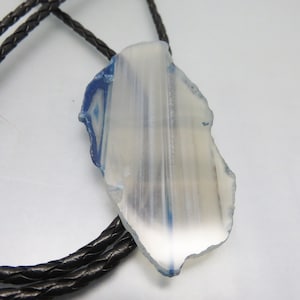 May include: A bolo tie featuring a translucent, irregularly shaped stone pendant with blue and white striations. The pendant is suspended from a braided black leather cord, creating a rustic and natural aesthetic.