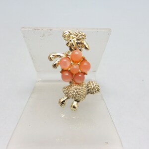 May include: Gold-tone brooch featuring a poodle with a pink rhinestone flower on its back.