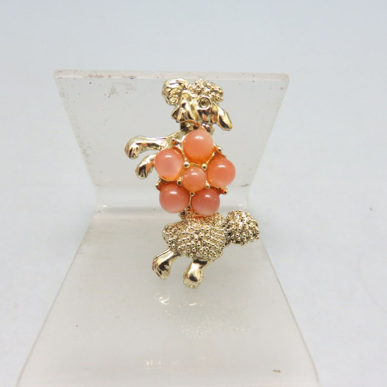 May include: Gold-tone brooch featuring a poodle with a pink rhinestone flower on its back.