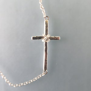 K&ouml;nnte beinhalten: Eine silberne Kettenkette mit einem silbernen Kreuzanh&auml;nger. Das Kreuz hat ein strukturiertes Design und ein gedrehtes Detail in der Mitte.