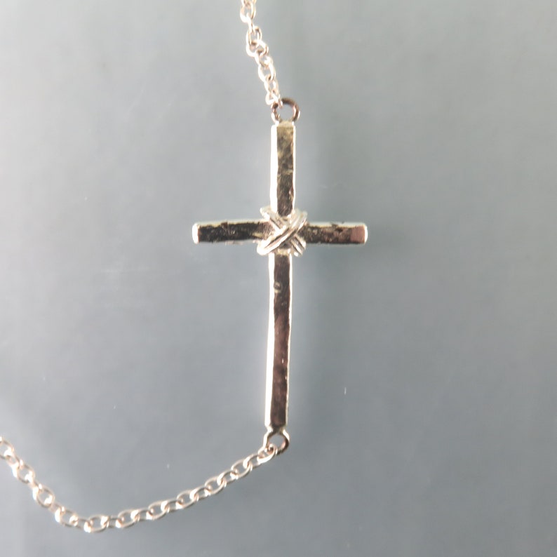 K&ouml;nnte beinhalten: Eine silberne Kettenkette mit einem silbernen Kreuzanh&auml;nger. Das Kreuz hat ein strukturiertes Design und ein gedrehtes Detail in der Mitte.