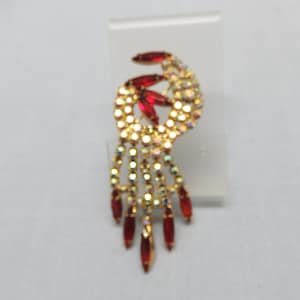 Pode incluir: Um broche dourado com um design pendurado. O broche apresenta um grupo de strass vermelhos e transparentes.