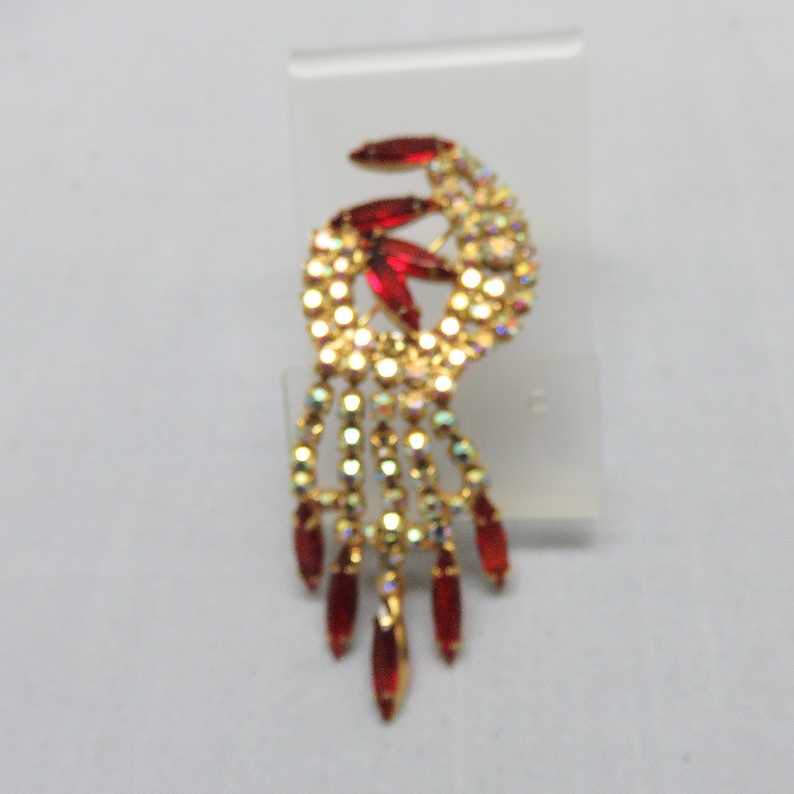 Pode incluir: Um broche dourado com um design pendurado. O broche apresenta um grupo de strass vermelhos e transparentes.