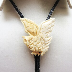 May include: A carved owl bolo tie with a black leather cord. The owl is made of white bone and has detailed carvings.