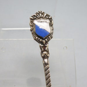 May include: Silver souvenir spoon with a blue and white enamel shield featuring the word "Zurich" on it.