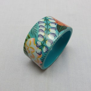 Op de afbeelding: Een turquoise bangle armband met een bloemenpatroon. Het patroon bevat witte, gele, roze, oranje en groene bloemen en bladeren.