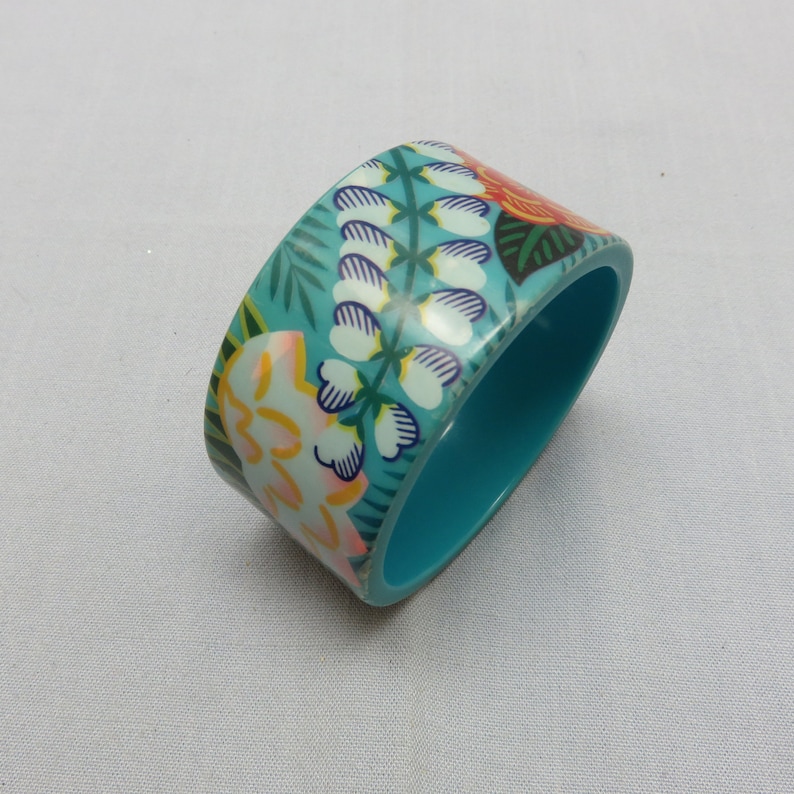 Op de afbeelding: Een turquoise bangle armband met een bloemenpatroon. Het patroon bevat witte, gele, roze, oranje en groene bloemen en bladeren.
