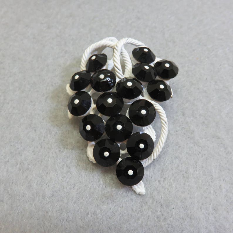 May include: A white metal brooch shaped like a bunch of grapes with black rhinestones. The rhinestones have a white dot in the center.