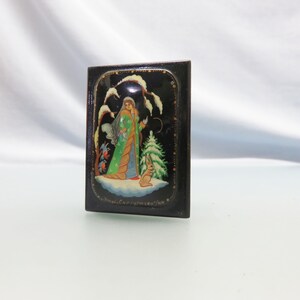 May include: A small black wooden box with a painted scene of a woman in a green and white dress standing in a snowy forest with a rabbit. The text "Хохлома С.Г.Русакова/1984" is written on the bottom of the box.