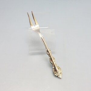 May include: A silver fork with an ornate handle. The fork has three tines and is designed for serving food.