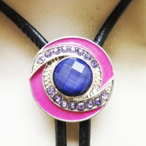 May include: A close-up of a bolo tie with a circular pendant. The pendant features a large, faceted blue stone at the center, surrounded by small purple gems and a bright pink enamel border. The tie has black cords.