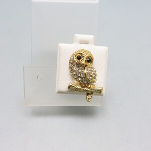 Pode incluir: Um broche de ouro em forma de coruja com strass transparentes e olhos pretos. A coruja está pousada em um galho.