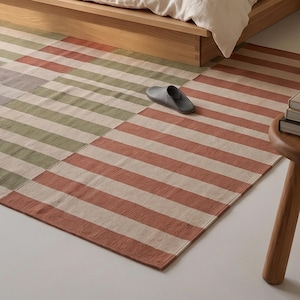 Op de afbeelding: Een rechthoekig tapijt met een gestreept patroon in tinten rood, groen en beige. Het tapijt ligt op een witte vloer in de buurt van een houten bedframe en een kleine houten bijzettafel. Een grijze slipper ligt op het tapijt.