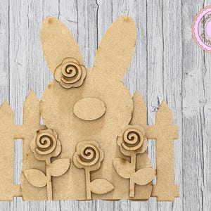 May include: A wooden Easter bunny cutout with a fence and rose accents. The bunny has large ears and an oval face. Three roses are placed in front of the bunny, and one rose is on the bunny's head. The background is a light wood grain.