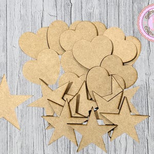 May include: A collection of unfinished wooden shapes, including hearts and stars, arranged on a light-colored wooden surface. The shapes vary in size and are ready for crafting or decoration. A circular logo with text is in the upper right corner.