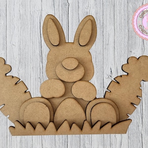 May include: A wooden Easter bunny with carrots and eggs. The bunny is in the center, with large ears and a round body. Carrots are on either side, and eggs are at the bottom. The design is made of layered wood.