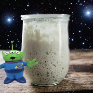 May include: A clear glass jar filled with a light-colored, bubbly substance, possibly sourdough starter, sits on a wooden surface. A green alien toy with three eyes and a blue spacesuit stands beside the jar, against a starry background.
