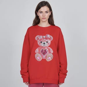 May include: A red sweatshirt featuring a sparkling teddy bear design. The bear has a pink bow, a heart-shaped gem, and is adorned with crystals. The sweatshirt is a solid red color.