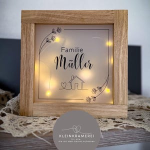 Può includere: Una scatola luminosa con cornice in legno con la scritta "Familie Müller". Il design include una casa e decorazioni floreali. La scatola è illuminata da piccole luci dai toni caldi, creando un'atmosfera accogliente. La cornice è in legno chiaro.