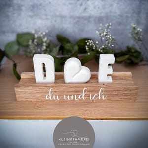 Könnte beinhalten: Ein dekoratives Holzstück mit den weißen Buchstaben "D", einem Herz und "E", angeordnet auf einem Holzsockel. Die Worte "du und ich" sind auf der Basis geschrieben. Der Hintergrund ist grau mit grüner Vegetation.