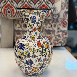 May include: A ceramic vase with a white base, adorned with a vibrant floral pattern in blue, yellow, red, and orange. The vase has a bulbous body and a flared neck with a black rim. The base has a light blue trim.