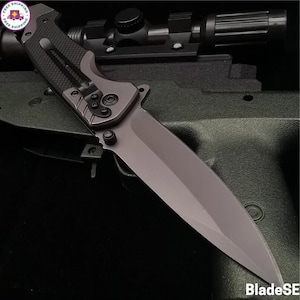 Puede incluir: Una navaja gris con mango texturizado y clip. La hoja es de color gris sólido. El cuchillo está sobre una superficie negra.