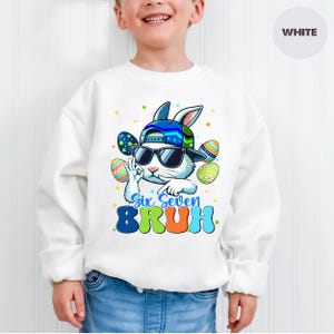 May include: White sweatshirt featuring a cartoon bunny wearing sunglasses and a baseball cap, surrounded by Easter eggs. The text "Six Seven BRUH" is printed in blue and orange. The sweatshirt is worn by a child.
